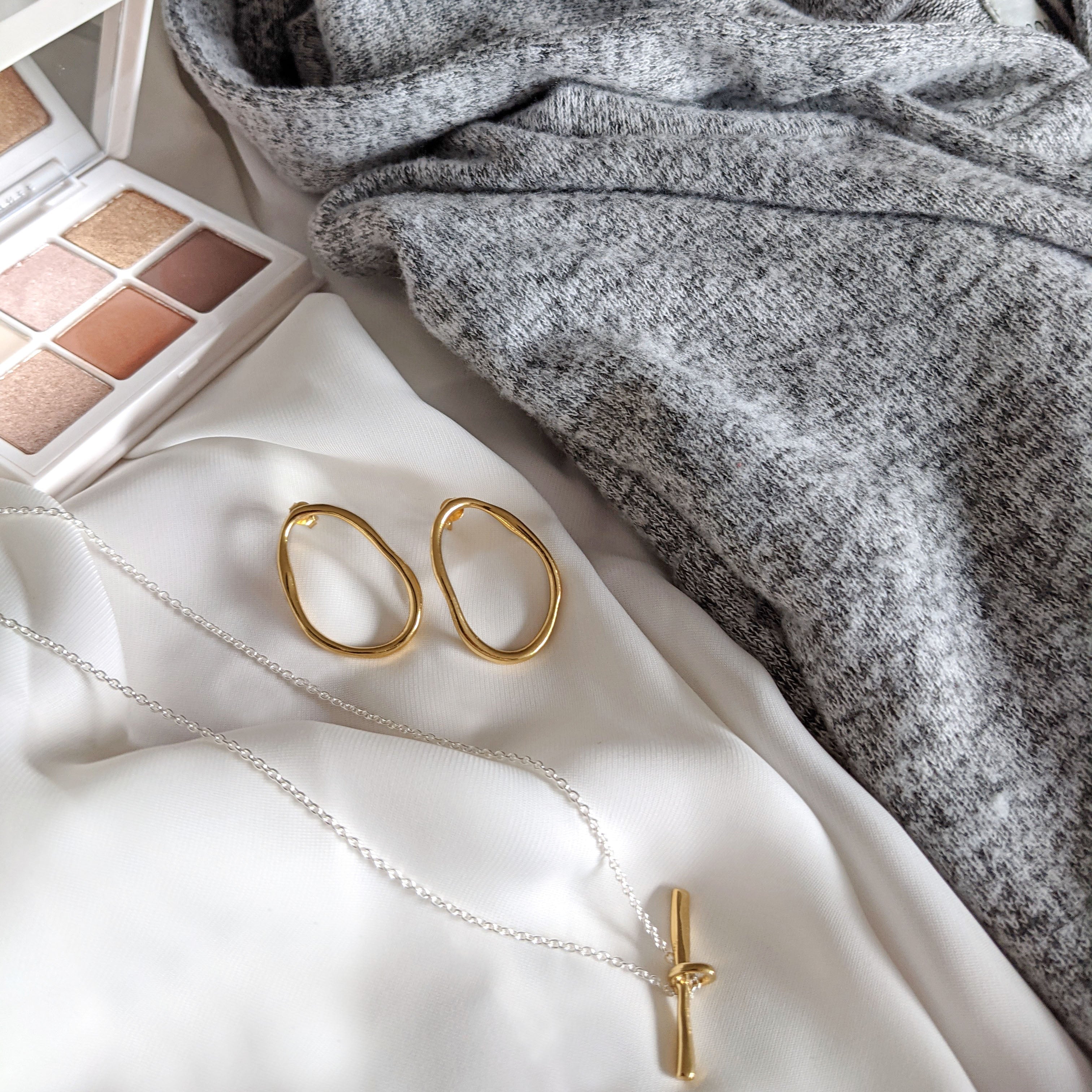 circle earrings and t-bar necklace lay on bed next to makeup pallet