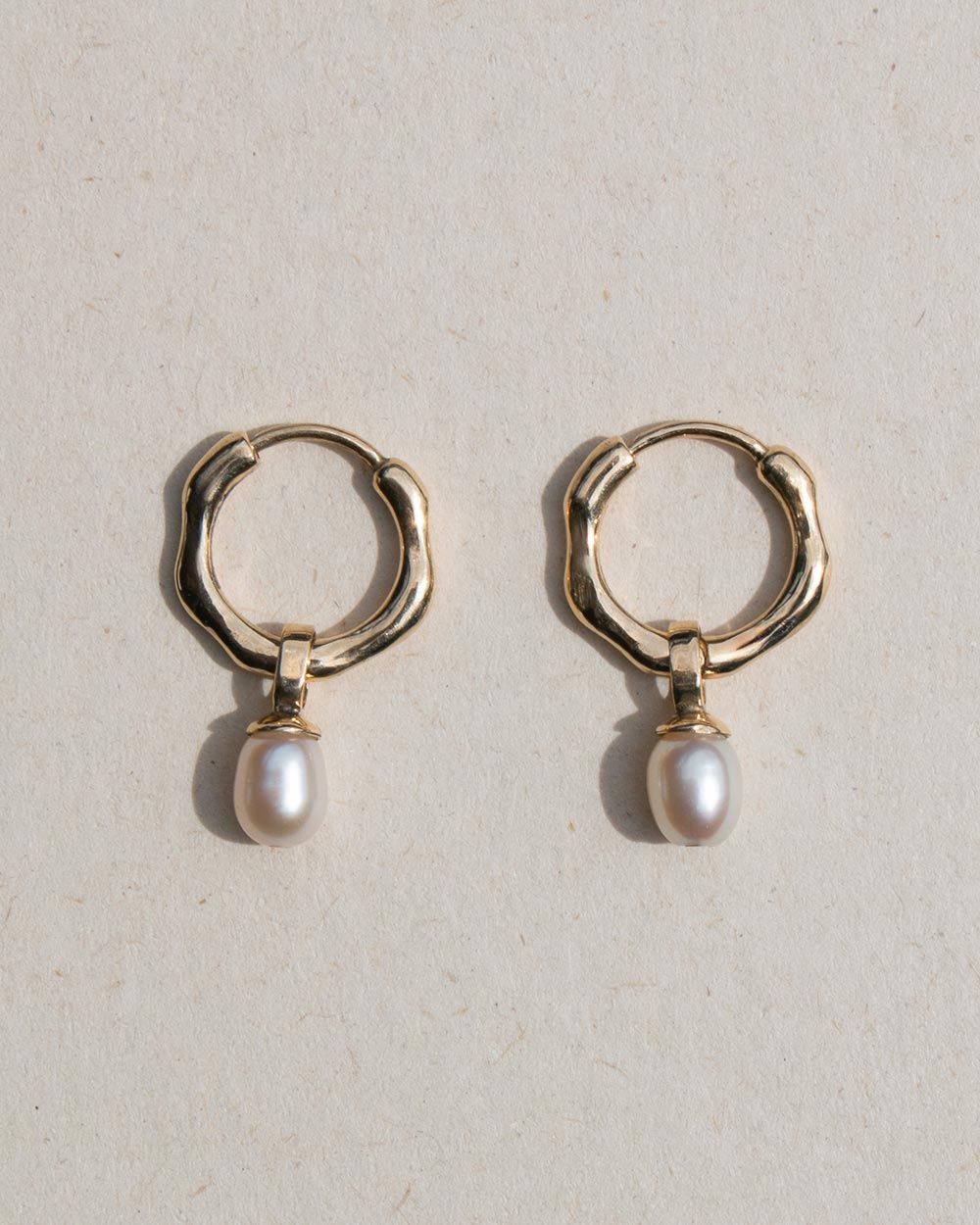 Front view of A pair of 9ct solid gold huggie hoop earrings with a dangling white baroque pearl, displayed on a textured beige background.