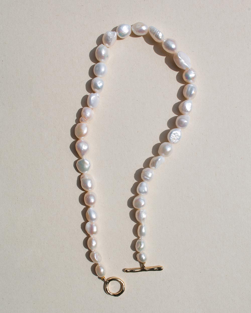 A baroque pearl necklace with a gold toggle clasp and ring, laid flat on a light beige background.