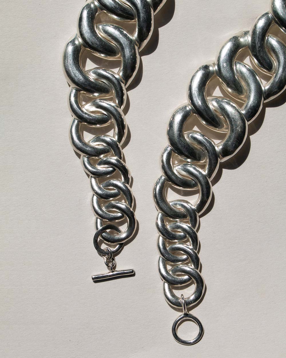 A close-up of a chunky silver chain bracelet with a toggle clasp and a hanging ring, photographed on a plain off-white background.