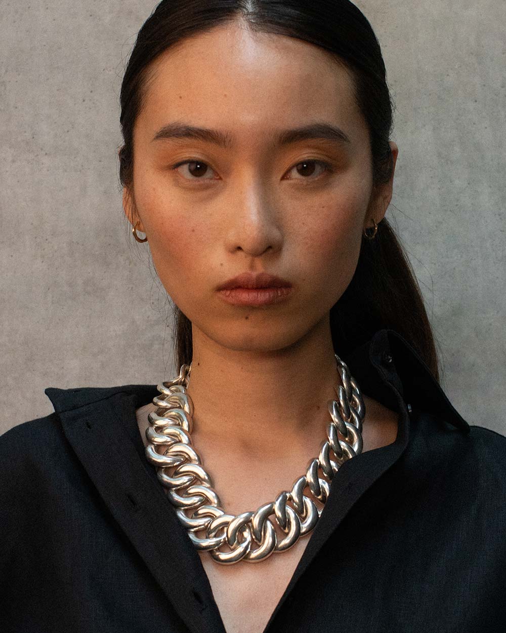 A close-up portrait of a model wearing a chunky silver chain necklace over a black collared shirt, set against a textured gray background.