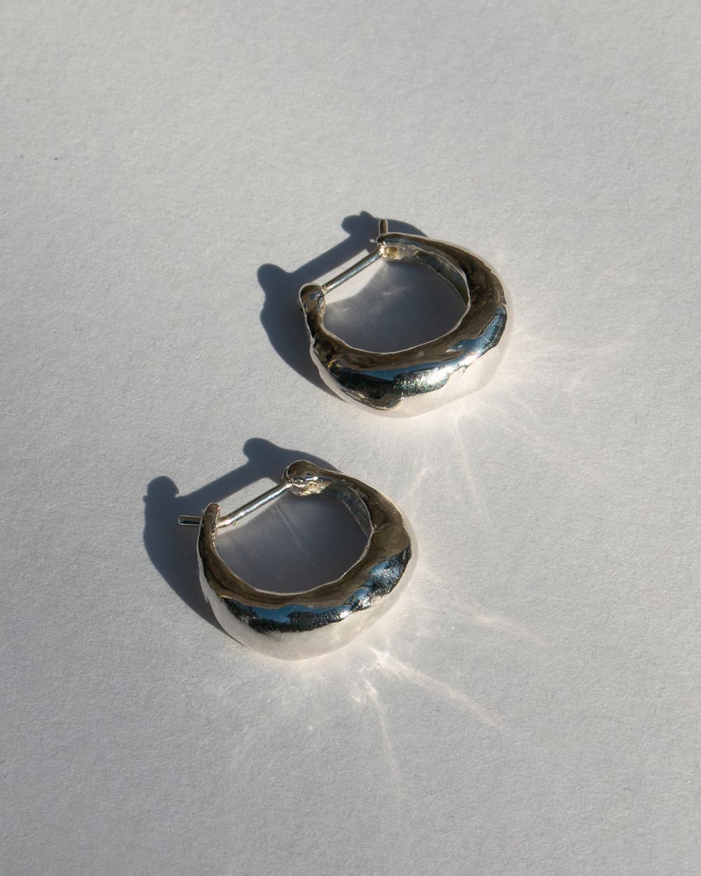 Top view of Sterling silver molten hoop earrings with a chunky organic texture, photographed on a light textured background with natural shadows.