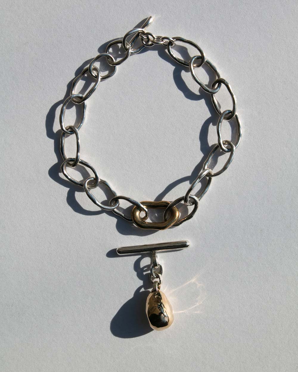 Sterling silver chain bracelet with a 9ct gold drop charm and toggle clasp, photographed on a light textured background with natural shadows. Charm detatched.