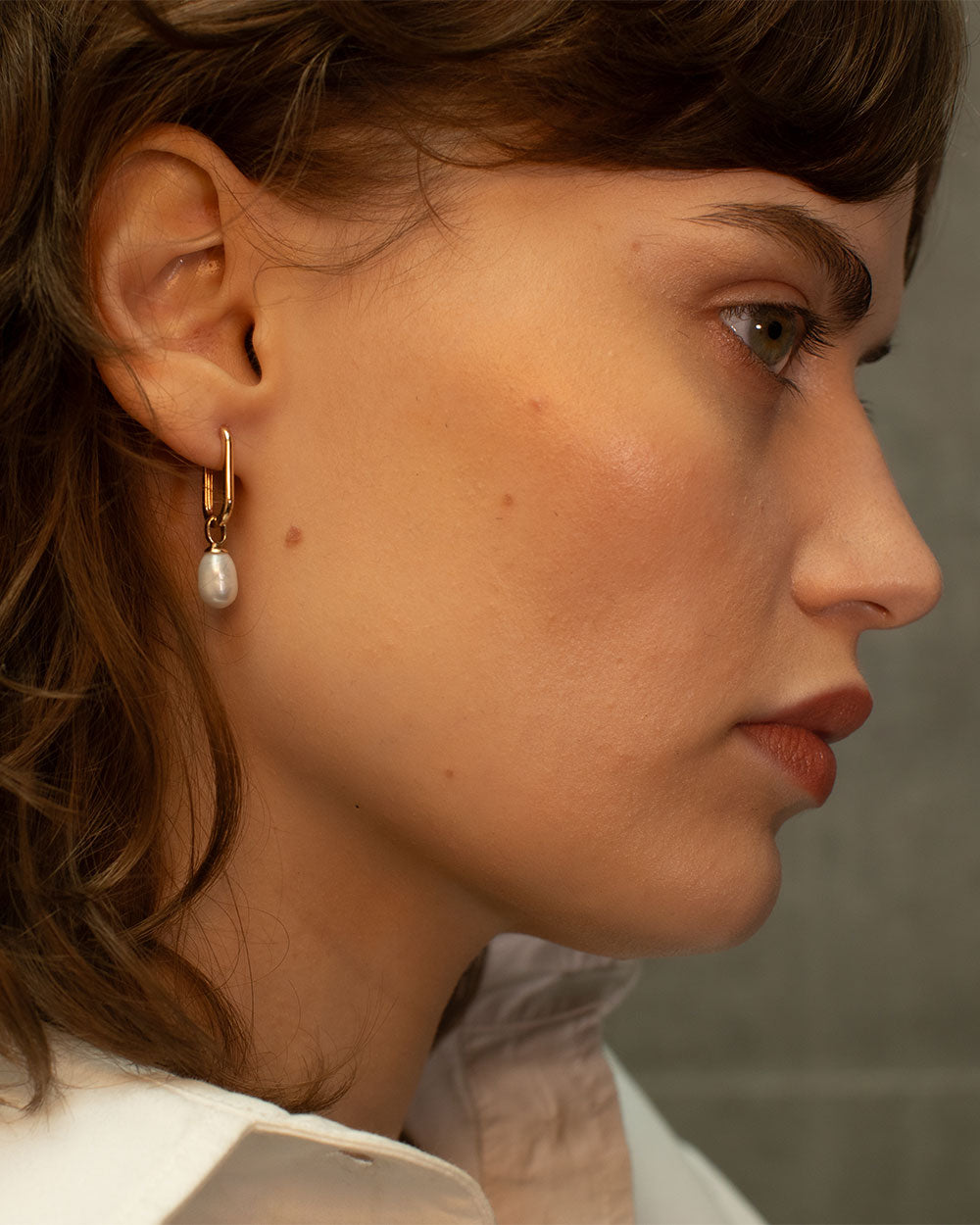 Close up side view of model wearing 9ct gold hoop earrings with pearl drop charms, photographed on a light textured background with natural shadows.