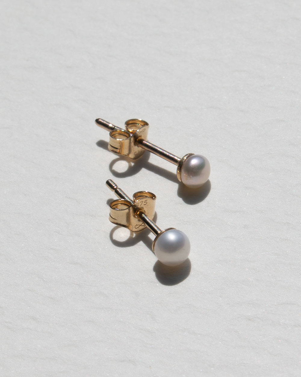 A pair of gold stud earrings with round white freshwater pearls, shown from a side angle on a textured off-white background.