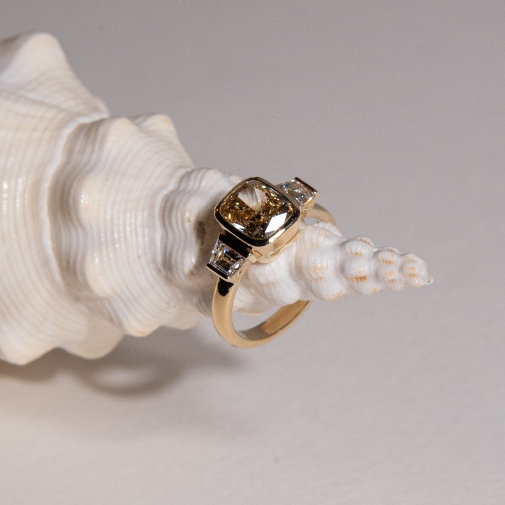 Bespoke champagne diamond engagement ring featuring a cushion-cut centre stone and baguette side diamonds in recycled gold, displayed on a white seashell.