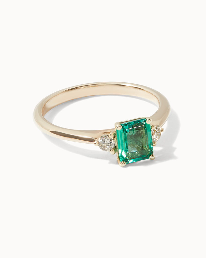 Solid gold engagement ring featuring a square emerald and two round cut white diamonds on the sides by Maya Magal London