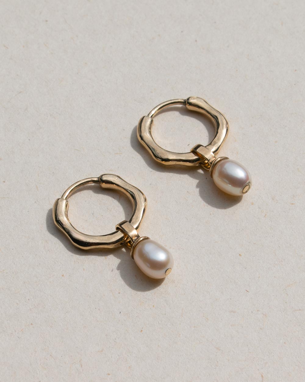 Side view of  A pair of 9ct solid gold huggie hoop earrings with a dangling white baroque pearl, displayed on a textured beige background.