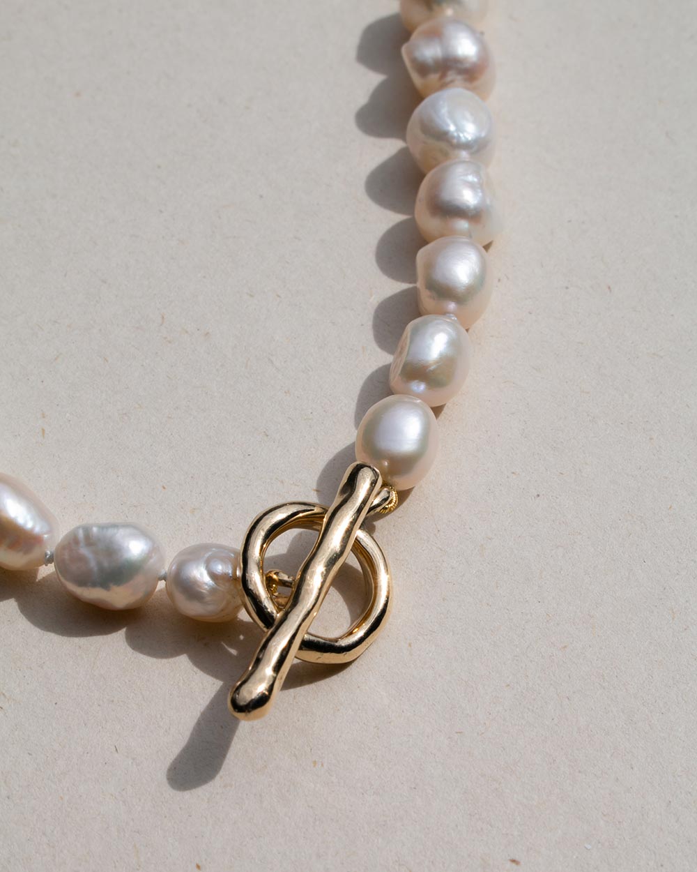 A close-up of a baroque pearl necklace with a polished gold toggle clasp, resting on a neutral beige surface.