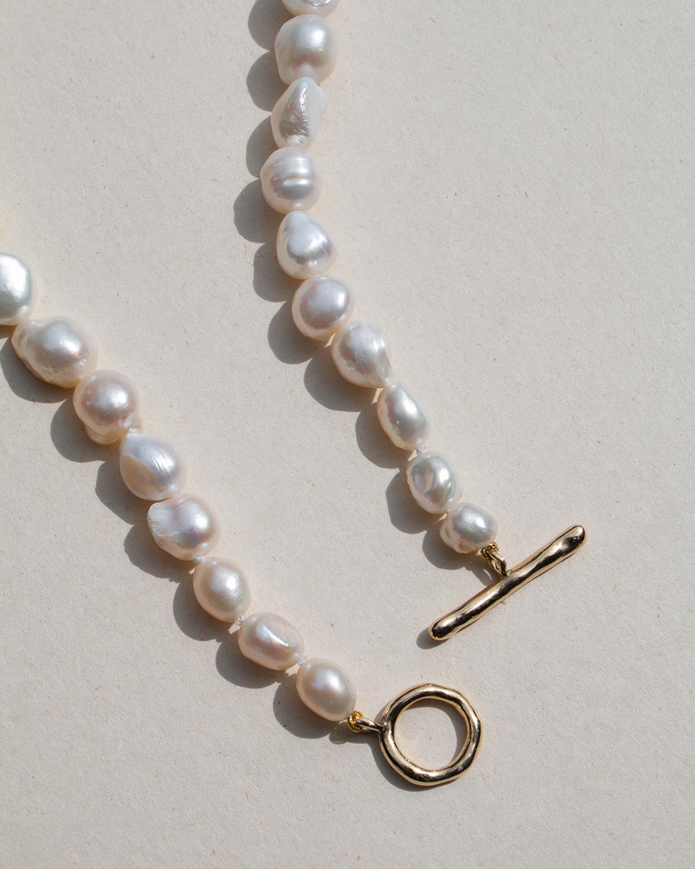 A close-up of a baroque pearl necklace with irregular white pearls and gold-toned hardware, including a toggle clasp and a round ring detail, laid on a light beige surface.