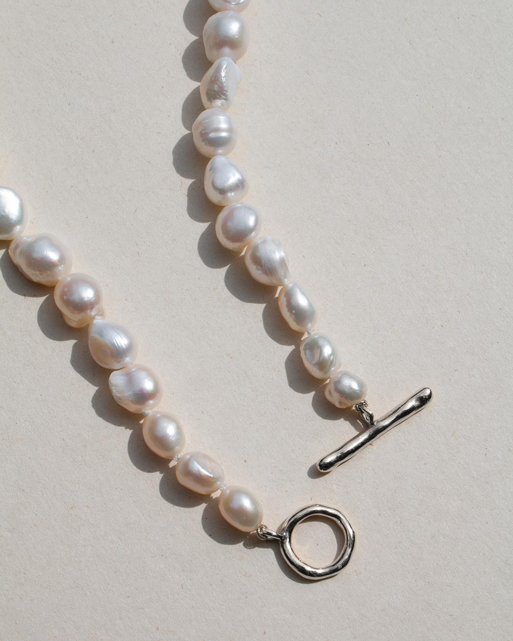 A close-up of a baroque pearl necklace with a silver toggle clasp and a textured silver ring detail, laid out on a warm beige background.