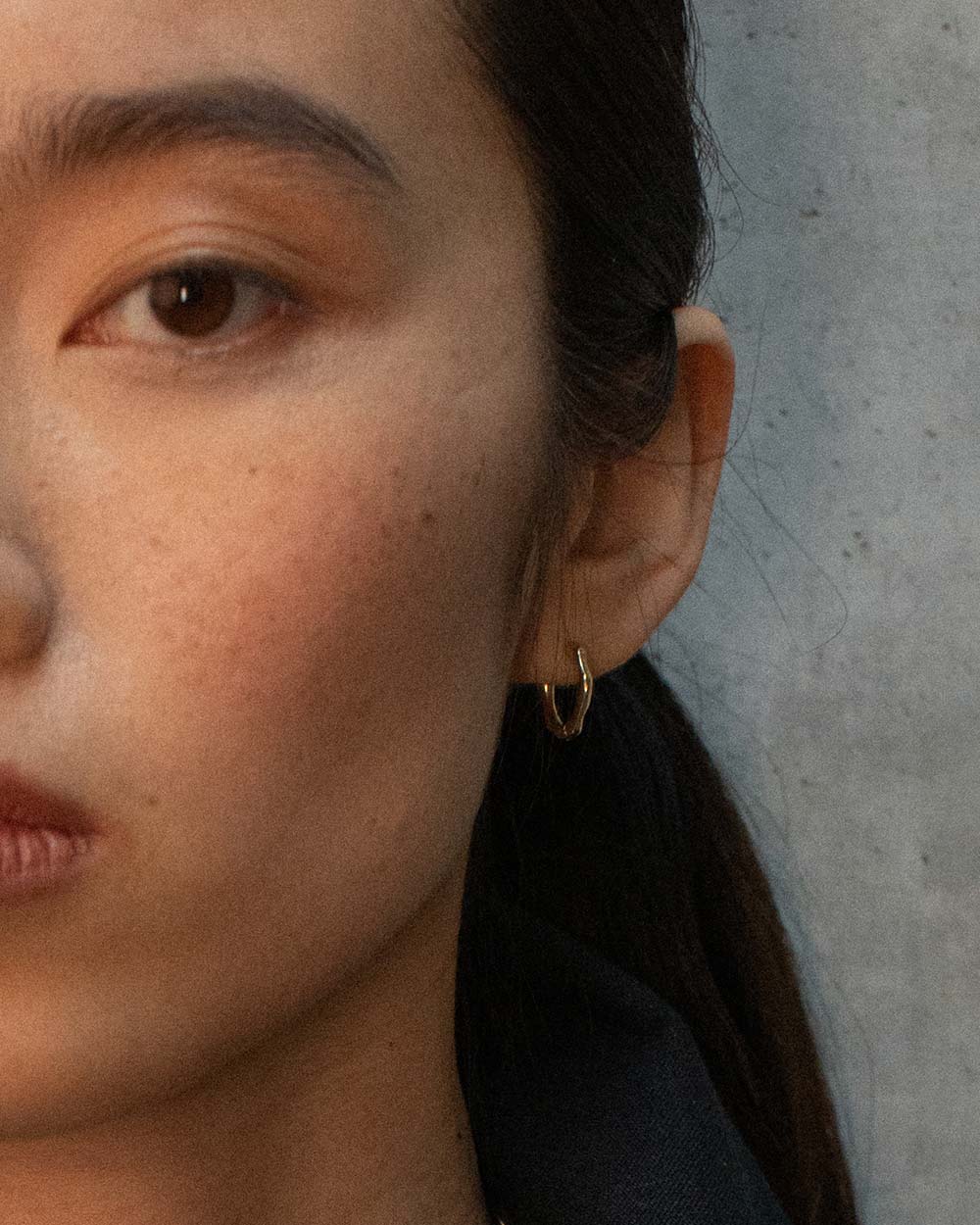 Close-up of a model's ear wearing a small gold hoop earring against a textured gray wall.