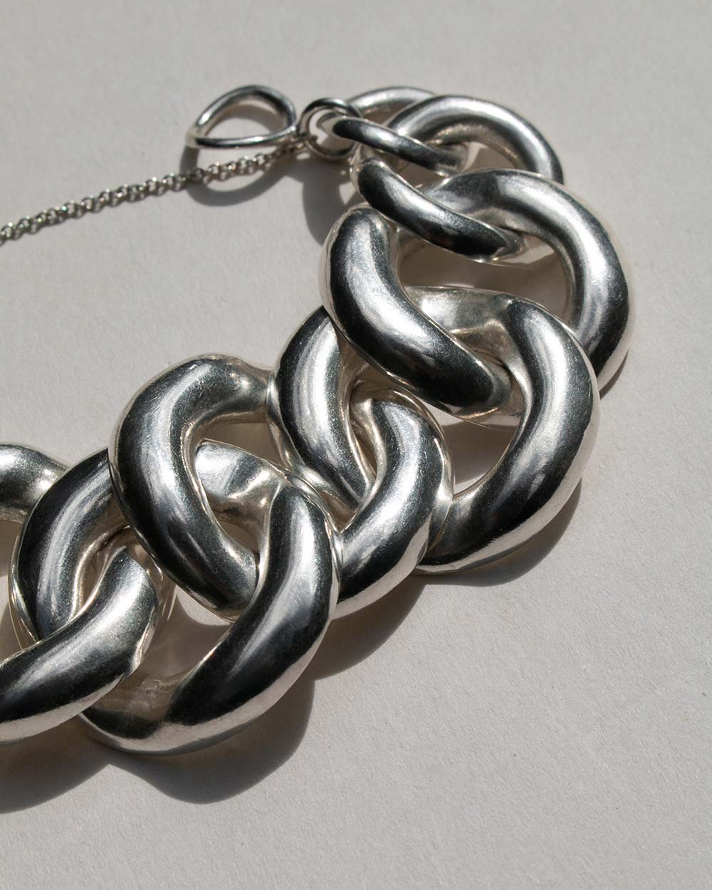 A close-up of a chunky silver chain link bracelet with thick, polished interlocking links and a delicate silver chain extender, shown on a light gray background.