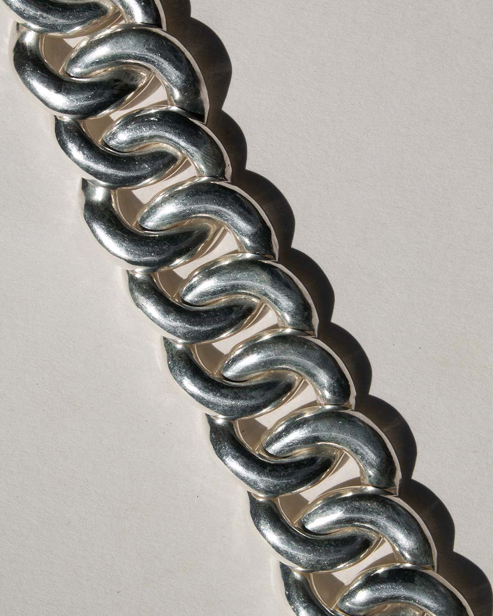 A close-up of a chunky silver curb link chain bracelet or belt, arranged diagonally on a light gray background.