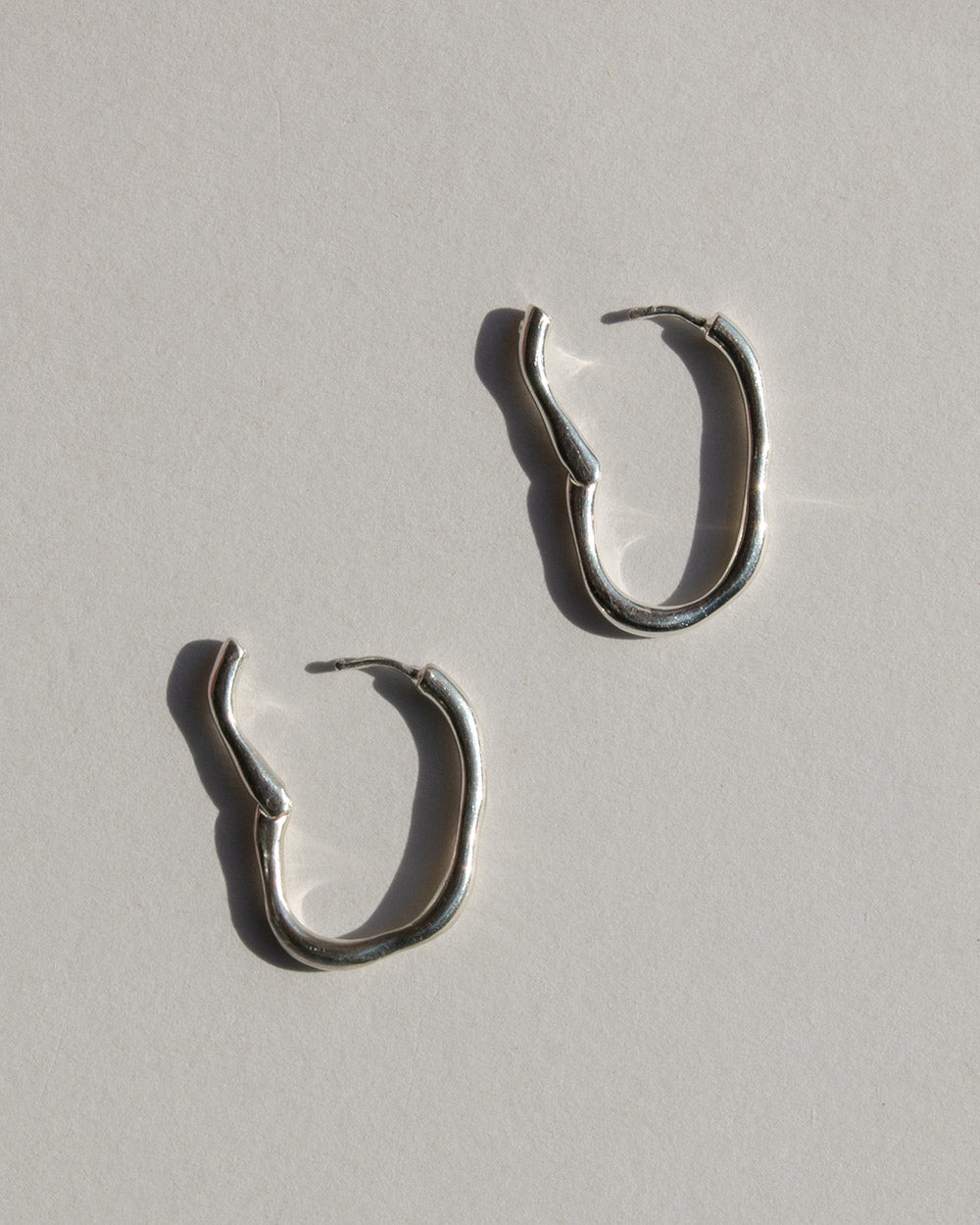 A pair of silver hoop earrings with a textured, organic wavy finish, displayed on a light gray textured surface with soft shadows.