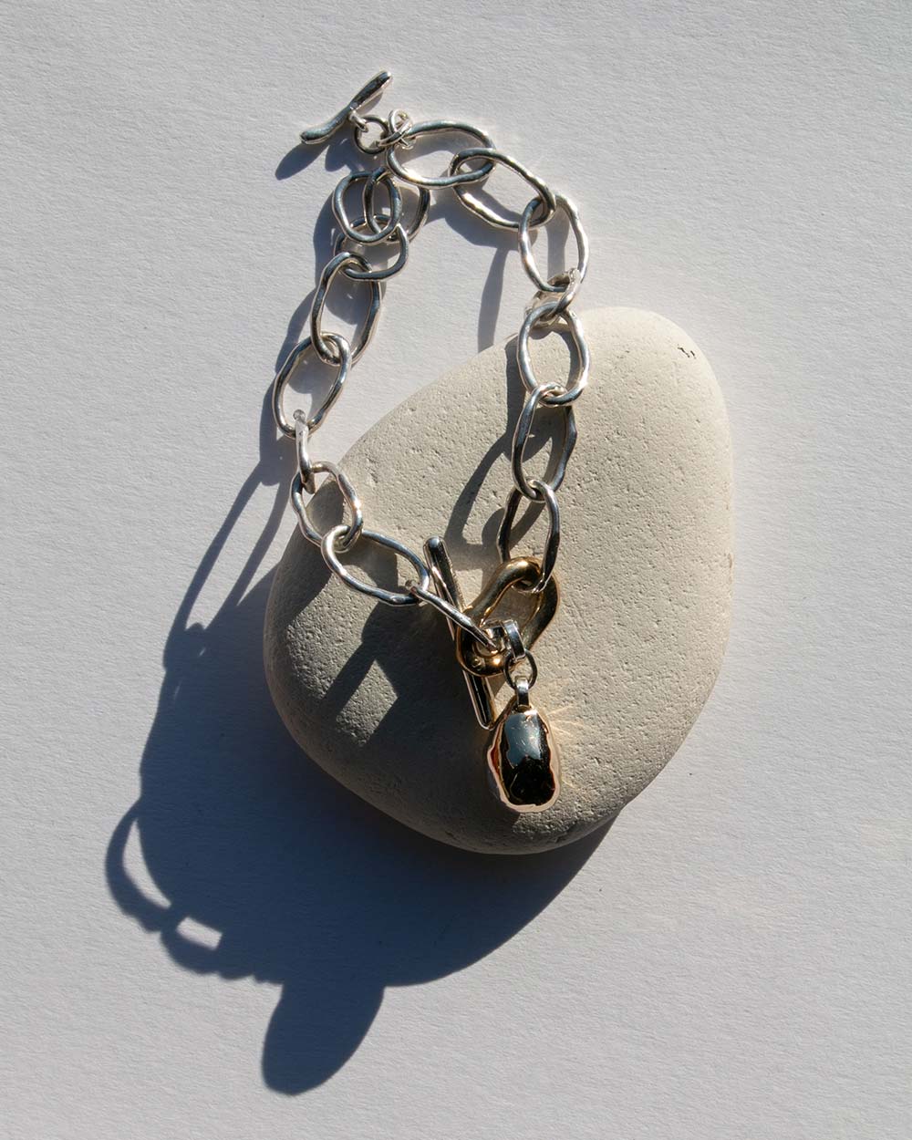 Sterling silver chain bracelet with a 9ct gold drop charm and toggle clasp, photographed on a light textured background with natural shadows. Displayed on rock