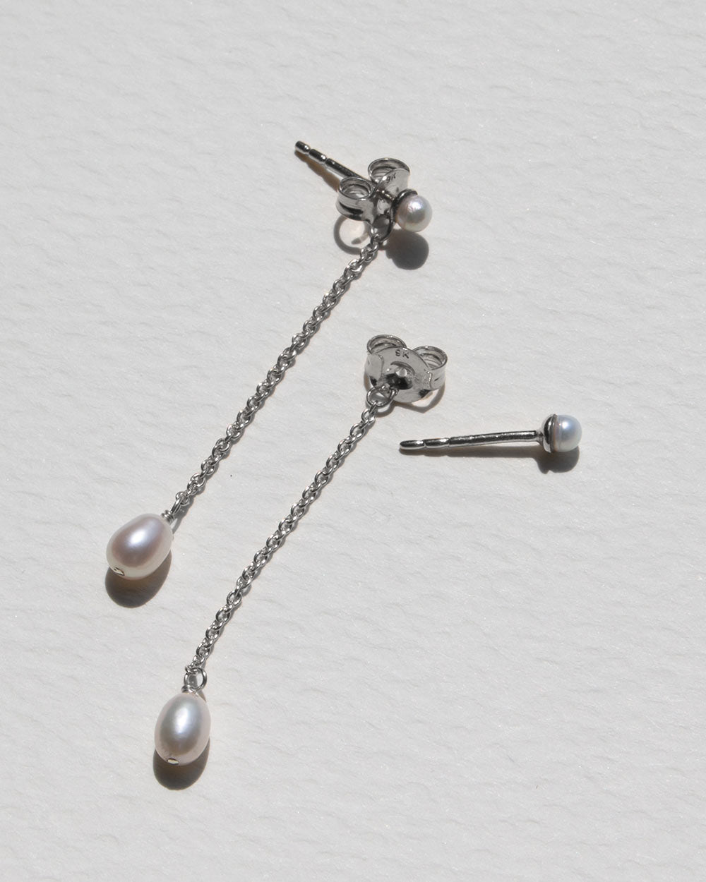 A pair of silver-tone dangle earrings with small oval white pearls on a textured off-white background. One earring is shown hanging from its back clasp, and the other is shown from the side.